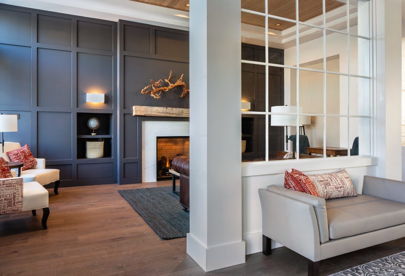 A modern living room with dark paneled walls, a lit fireplace, white armchairs with red patterned pillows, a gray sofa, wood flooring, and a partition with large square glass panes.