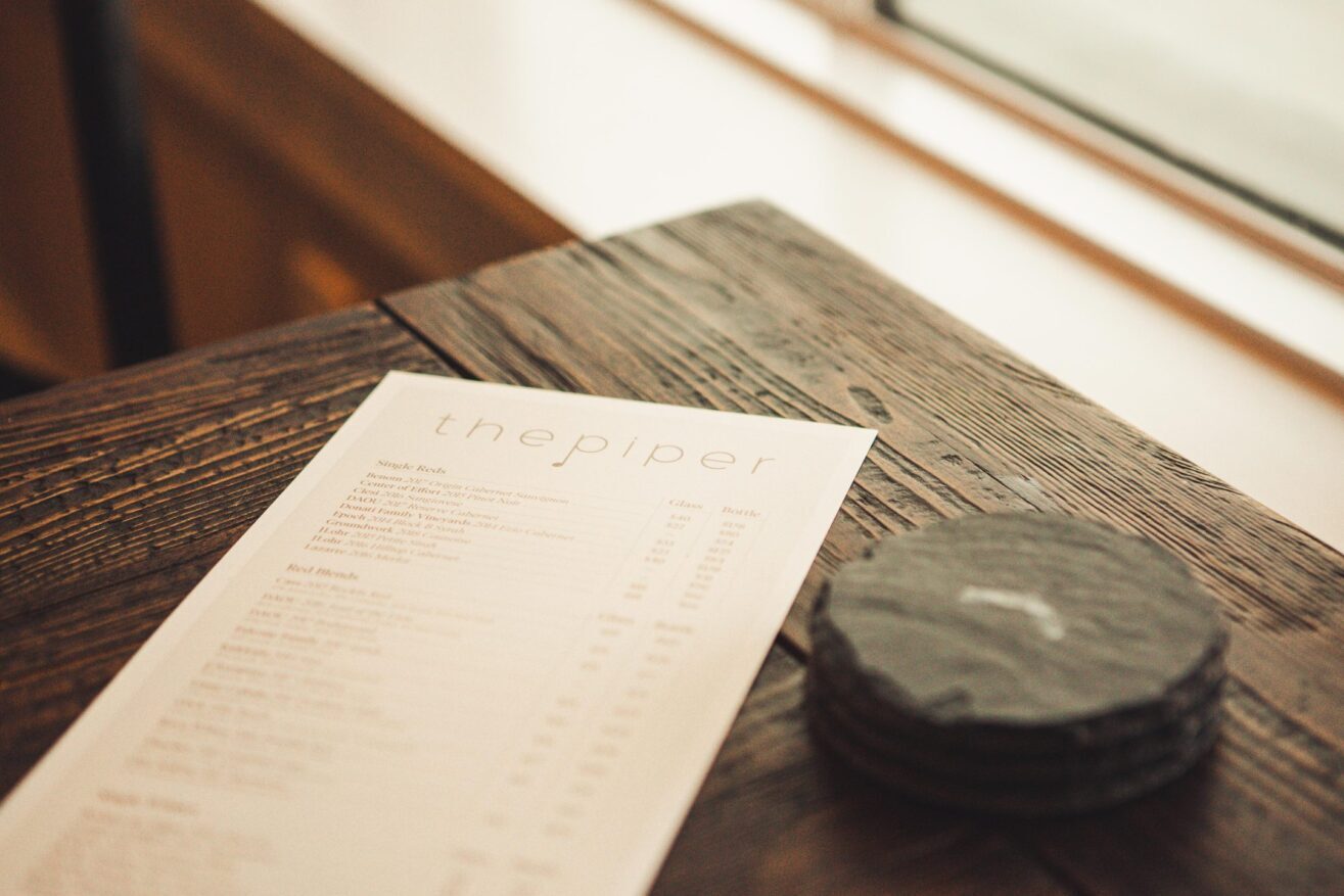 A restaurant menu titled the piper lies on a rustic wooden table next to a small stack of round, dark drink coasters by a window.