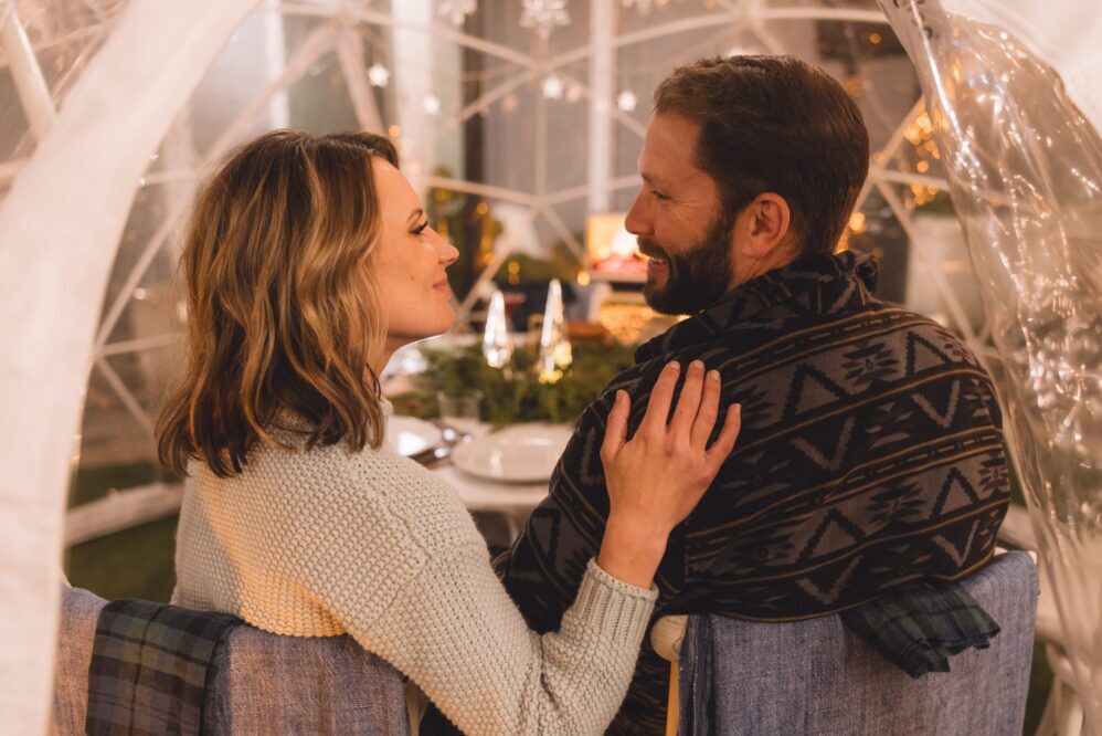 A couple sits close together inside a cozy, decorated dome during Holidays at Paso Robles Inn, smiling at each other with a festive table set for dinner in the background. The woman has her hand affectionately on the man's shoulder.