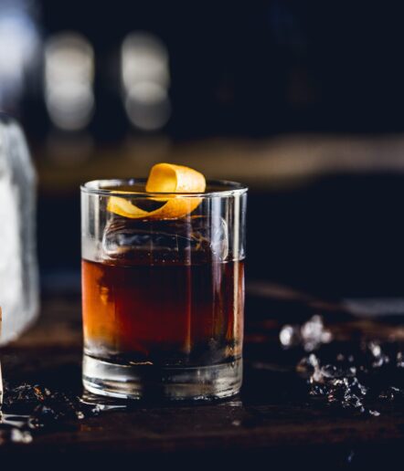A glass of dark cocktail with a large ice cube and an orange twist garnish sits on a wooden surface, with scattered ice pieces and a dark, blurred background.
