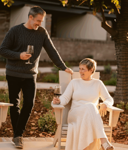 A man and woman enjoy glasses of red wine outdoors. The man stands smiling, while the woman sits in a wooden chair, also smiling. Both are dressed warmly, surrounded by sunlight and a garden setting.