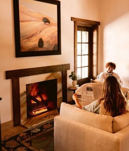 Two people relax in a cozy living room; one reads a newspaper on a cream sofa, the other sits in an armchair by a window. A lit fireplace and landscape painting create a warm, inviting atmosphere.