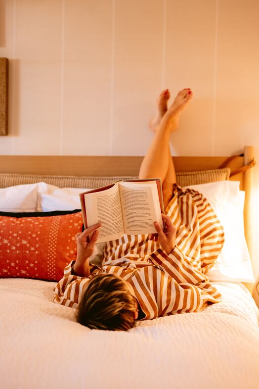 A person in a striped dress lies on a bed with their legs resting up against the wall, holding and reading an open book. Cozy pillows are arranged behind them. The room is softly lit.