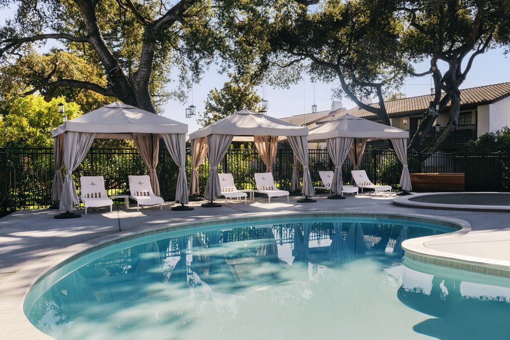 Sunny outdoor pool with clear water, surrounded by white lounge chairs and four cabanas with curtains, set beneath large leafy trees and near a building with a tiled roof.