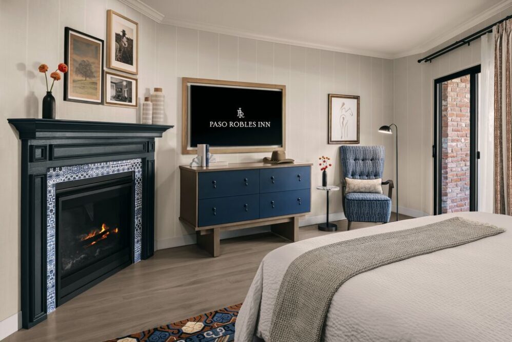 A cozy hotel room with a bed, fireplace, dresser with a TV displaying Paso Robles Inn, an armchair with a lamp, framed wall art, and a door leading to a brick patio. The decor is modern and inviting.
