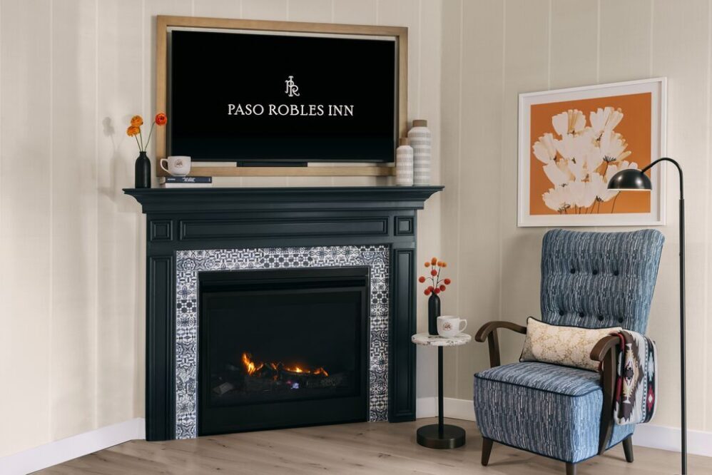 A cozy corner with a blue armchair, side table, and floor lamp beside a black fireplace. A TV above the mantel displays “Paso Robles Inn,” and there’s modern art and simple, colorful decor in the room.