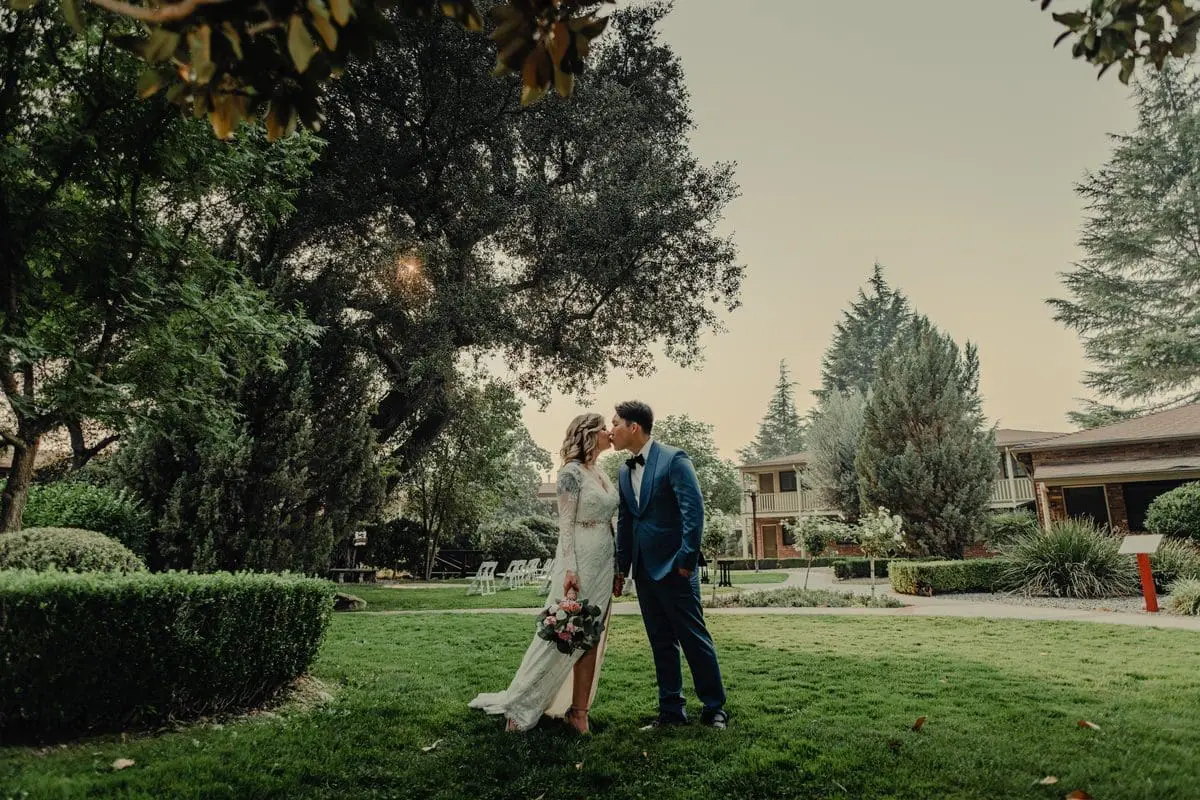 Bride and groom embracing in a sunlit outdoor garden, surrounded by vibrant flowers and greenery, with the bride holding a bouquet and the groom in a dark suit.