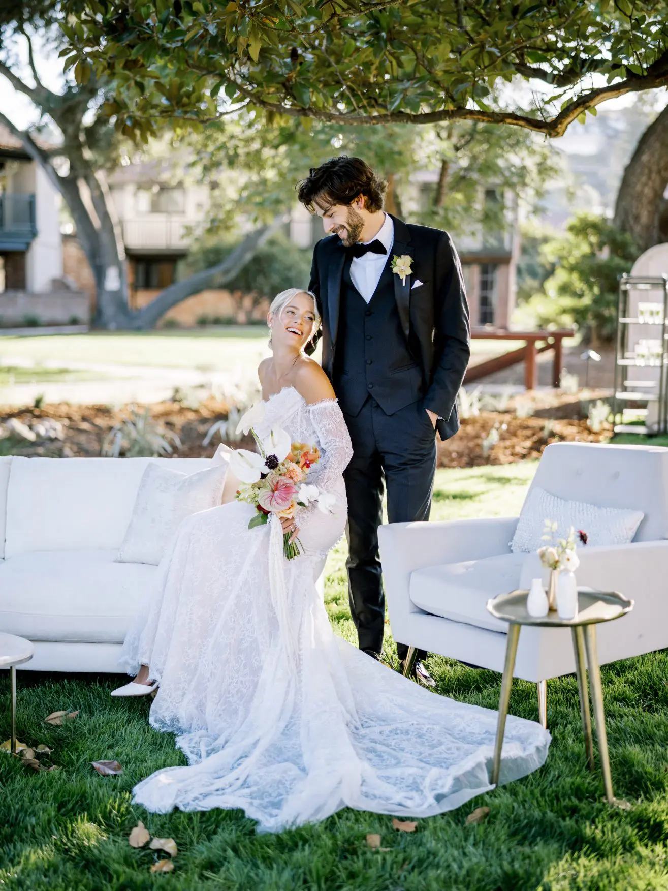 A bride in a white wedding dress sitting on a white sofa with a groom in a black suite smiling down at her in a garden.