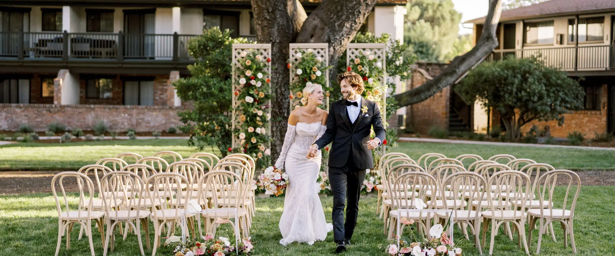 A bride in a white dress holding hands with the groom in a black suite running down the aisle of wooden wedding ceremony chairs in a garden.